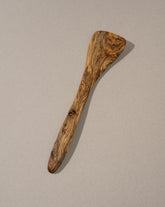 A single natural olive wood spatula with a smooth finish and characteristic wood grain patterns, displayed against a neutral background.